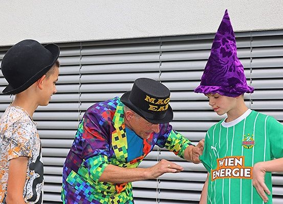 Ein Mann in einem farbenfrohen Outfit führt einen Zaubertrick vor zwei Jungen vor, einer trägt einen lila Hut und der andere ein grünes Trikot.