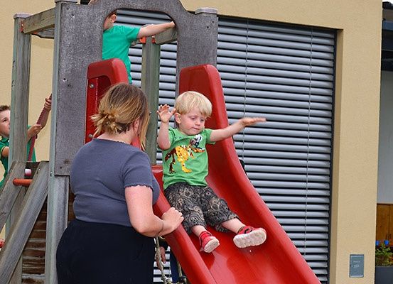 Mehrere Kinder spielen auf einem Spielplatz, ein Kind rutscht auf einer roten Rutsche, während eine Erwachsene zuschaut. Dahinter steht ein Gebäude mit Jalousien.
