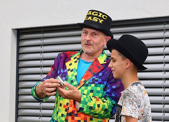 Ein Zauberer in einem bunten Kostüm und Hut zeigt einem jungen Jungen einen Trick. Der Junge trägt einen schwarzen Hut und ein weißes T-Shirt. Sie stehen vor einer weißen Wand mit Jalousien.