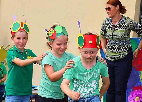 Eine Gruppe von Kindern mit Raupen-Kopfbändern und grünen T-Shirts. Eine Frau steht hinter ihnen, trägt eine Sonnenbrille und eine gemusterte Bluse.