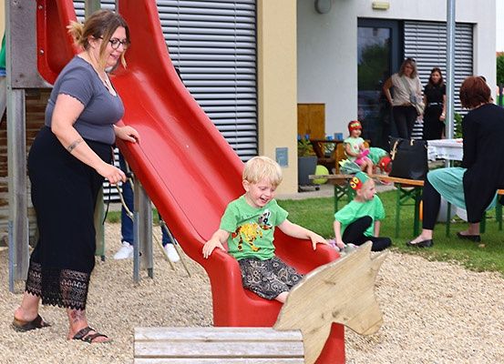 Eine Frau hilft einem jungen Jungen, eine rote Rutsche auf einem Spielplatz hinunterzurutschen. In der Nähe spielen andere Kinder auf dem Gras.