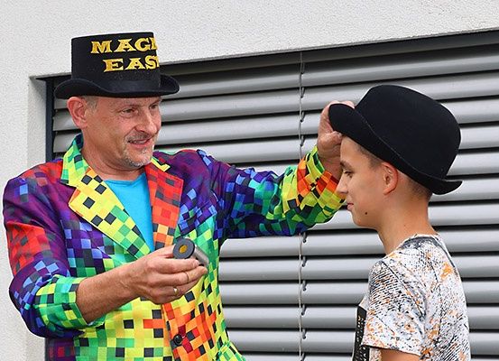 Ein Mann in einem farbenfrohen, mehrfarbigen Anzug und einem Hut mit der Aufschrift 'MAGIC EASY' steht neben einem jungen Jungen in einem weißen T-Shirt, beide betrachten ein Objekt, das der Mann in der Hand hält. Sie stehen vor einem Gebäude mit horizontalen Jalousien.
