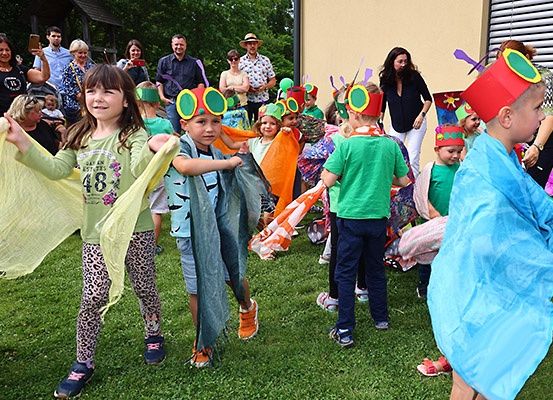 Kinder, die wie Käfer gekleidet sind, ziehen auf einem Rasen vorbei. Erwachsene beobachten im Hintergrund. Die Kinder tragen bunte Mützen und tragen leichte Stoffe.
