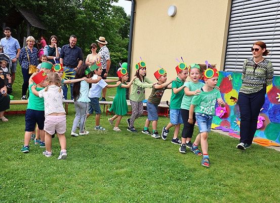 Eine Gruppe von Kindern mit grünen Mützen und Schmetterlingsflügeln tanzt auf einer Wiese. Dahinter schmückt eine bunte Wandmalerei die Wand. Mehrere Erwachsene und Kinder beobachten.