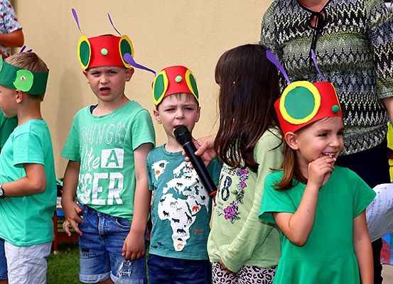 Eine Gruppe von Kindern, die bunte Käfermützen tragen, steht in einer Reihe, einer hält ein Mikrofon und ein anderer ein Stück Papier. Sie tragen alle grüne T-Shirts und scheinen an einer Veranstaltung teilzunehmen.