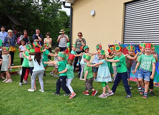 Kinder, die grüne Hüte tragen, tanzen im Kreis auf einer Rasenfläche, mit Erwachsenen und einem bunten Wandbild im Hintergrund.