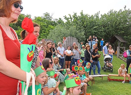 Eine Gruppe von Kindern und Erwachsenen versammelt sich in einem Park. Sie tragen Raupen-Kopfbänder. Eine Frau hält eine rote Papiertüte. Es gibt einen Kinderwagen und eine Holzbank in der Gegend.