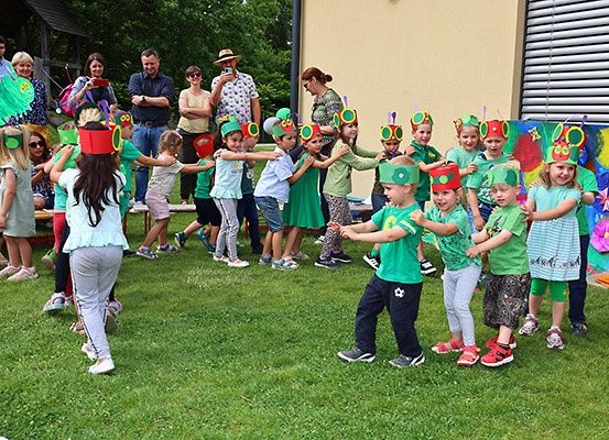 Eine Gruppe von Kindern in Kostümen tanzt auf einem Rasen, während Erwachsene im Hintergrund zusehen. Die Kinder tragen rote und grüne Hüte mit Antennen.