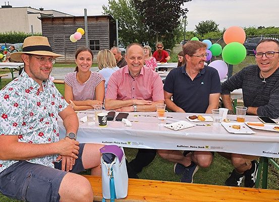 Eine Gruppe von Menschen sitzt um einen Tisch in einer Außenumgebung, lächelt und posiert für ein Foto. Bunte Luftballons und ein Zaun sind im Hintergrund zu sehen.