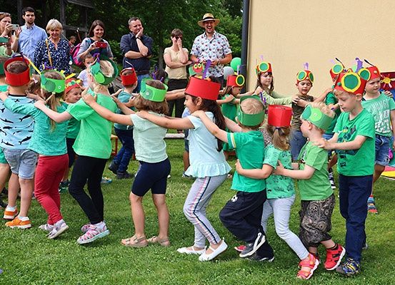 Eine Gruppe von Kindern, die grüne Hemden und Kopfbänder tragen, führt draußen auf einem Rasenfeld einen Tanz auf. Einige von ihnen tragen Turnschuhe, andere Sandalen. Hinter ihnen stehen einige Erwachsene und beobachten sie. Einige der Erwachsenen machen Fotos.