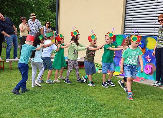 Eine Gruppe von Kindern in grünen Kleidern und roten Hüten tanzt draußen auf dem Gras. Erwachsene stehen hinter ihnen. Eine Frau lächelt. Eine Wand mit bunter Kunst ist hinter den Kindern.