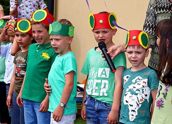 Mehrere Kinder, die bunte Raupenkopfbedeckungen tragen, stehen in einer Reihe, eines hält ein Mikrofon, wahrscheinlich während einer Aufführung oder Veranstaltung.