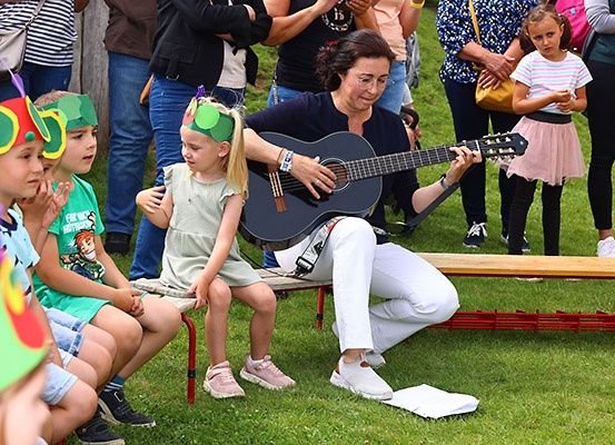 Eine erwachsene Frau spielt Gitarre im Park, während Kinder in Kostümen zusehen. Ein junges Mädchen steht hinter ihr.