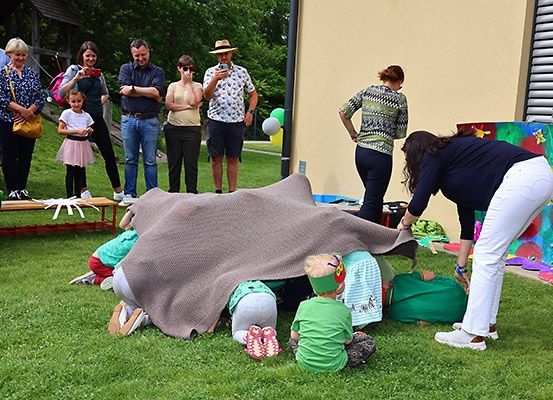 Mehrere Erwachsene und Kinder versammeln sich in einem Park. Einige Kinder spielen unter einer Decke, während andere zuschauen. Es gibt Luftballons und eine Wandmalerei an der Wand.
