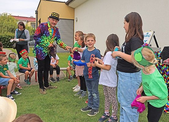 Ein bunt gekleideter Mann unterhält eine Gruppe von Kindern im Freien. Sie halten bunte Gegenstände in den Händen und beobachten ihn aufmerksam.