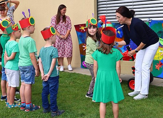 Eine Gruppe von Kindern, die Mückenkappen tragen, möglicherweise für ein Theaterstück, mit einer erwachsenen Frau, die ein Mikrofon hält, und einem bunten Gemälde dahinter.