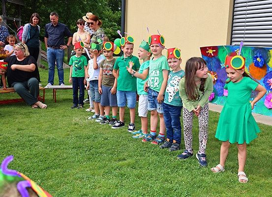 Mehrere Kinder mit Hutdekorationen stehen in einer Reihe, während Erwachsene an einem sonnigen Tag zusehen.