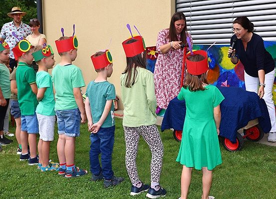 Kinder in grünen Shirts und roten Hüten versammeln sich auf einem Rasen für eine Aufführung. Eine Frau hält ein Mikrofon, und ein bunter Hintergrund ist hinter ihr.