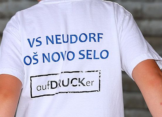 Eine Person trägt ein weißes T-Shirt mit blauem Text, der 'VS NEUDORF OS NOVO SELO' und schwarzen Text 'aufDRUCKer' auf der linken Seite steht.