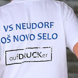 Eine Person trägt ein weißes T-Shirt mit blauem Text, der 'VS NEUDORF OS NOVO SELO' und schwarzen Text 'aufDRUCKer' auf der linken Seite steht.