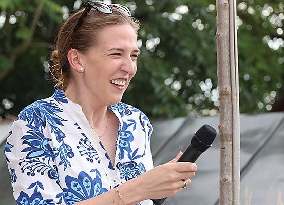 Eine Frau in einem blau-weißen Blumenhemd hält ein Mikrofon und lächelt, möglicherweise bei einer Veranstaltung im Freien.