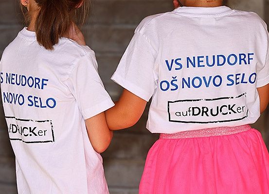 Zwei Personen tragen weiße T-Shirts mit blauem Text und stehen eng beieinander. Das linke Shirt sagt 'S Neudorf Novo Selo'. Das rechte Shirt liest 'VS Neudorf Os Novo Selo'. Beide Shirts haben ein Logo 'aufdrucker'.