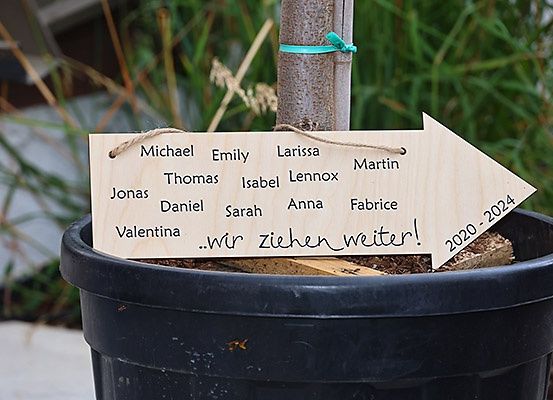 Ein Holzschild mit Namen wie Michael, Emily, Jonas und anderen ist an einem Baum in einem Topf befestigt. Das Schild hat eine handgeschriebene Nachricht, die 'Wir gehen weiter!' und einen Datumsbereich von 2020-2024 besagt.
