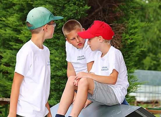 Drei Jungen in weißen T-Shirts und Mützen stehen auf einer grauen Plattform. Ein Junge sitzt mit überkreuzten Beinen. Hinter ihnen sind grüne Büsche.