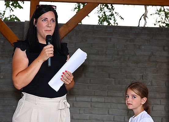 Eine Frau in Schwarz hält ein Mikrofon und Papier und spricht im Freien. Ein junges Mädchen in Weiß schaut zu. Hinter ihnen ist eine Ziegelmauer zu sehen.