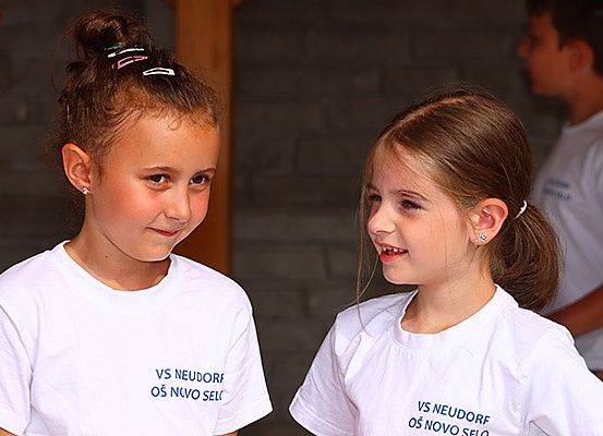 Zwei junge Mädchen in identischen weißen T-Shirts mit der Aufschrift 'VS NEUDORF OS NOVO SELO' lächeln und schauen sich an. Dahinter ist ein junger Junge teilweise zu sehen.