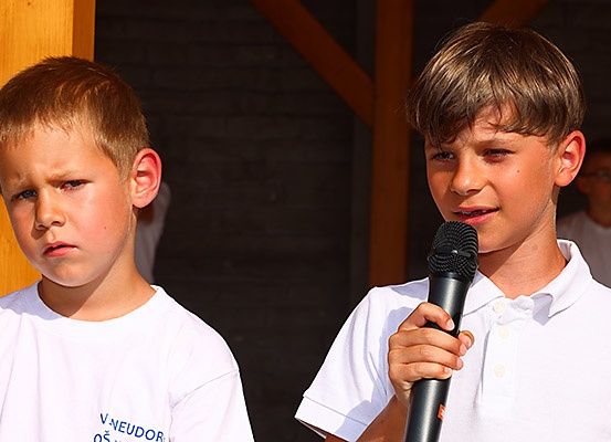 Zwei Jungen in weißen Poloshirts stehen im Freien. Ein Junge hält ein Mikrofon und scheint zu sprechen, während der andere Junge zusieht. Eine Person steht im Hintergrund.