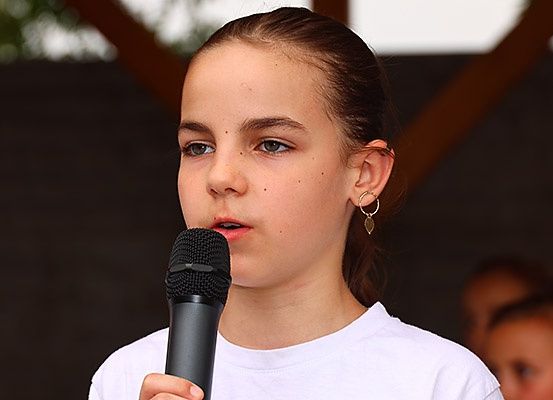 Ein junges Mädchen hält ein Mikrofon und scheint bei einer Veranstaltung im Freien zu sprechen. Sie trägt ein weißes T-Shirt. Im Hintergrund befinden sich verschwommene andere Personen.