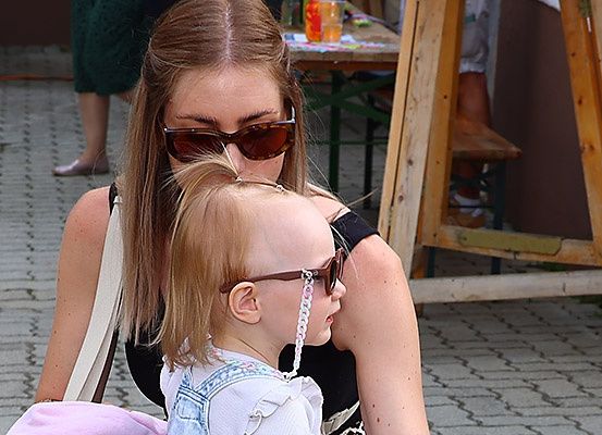 Eine Frau mit Sonnenbrille hält ein Kleinkind in einer Jeans-Latzhose, beide tragen Sonnenbrillen. Sie sind auf einem gepflasterten Weg, mit einem verschwommenen Tisch und einer Bank im Hintergrund.