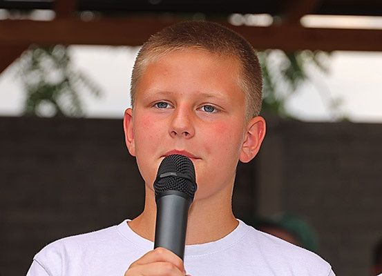 Ein junger Junge mit kurzen blonden Haaren hält ein Mikrofon und singt wahrscheinlich bei einer Veranstaltung im Freien.