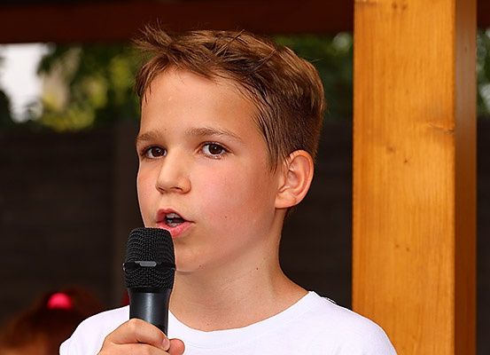 Ein junger Junge in einem weißen T-Shirt spricht in ein Mikrofon, wahrscheinlich bei einer Veranstaltung im Freien, mit einem hölzernen Pfosten hinter ihm.