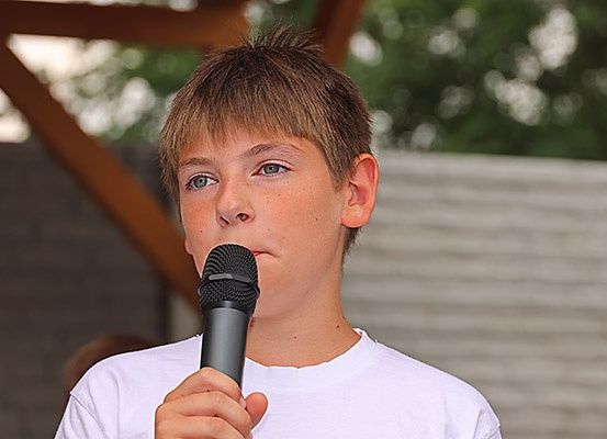 Ein Junge mit braunen Haaren spricht in ein Mikrofon. Er trägt ein weißes T-Shirt und sieht ernst aus.