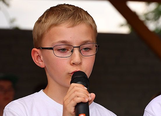 Ein junger Junge mit Brille hält ein Mikrofon und tritt wahrscheinlich bei einer Outdoor-Veranstaltung auf. Er trägt ein weißes T-Shirt. Dahinter ist eine verschwommene Figur in einem Hut zu sehen.