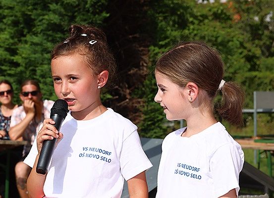 Zwei Mädchen mit Pferdeschwänzen, beide tragen weiße T-Shirts mit blauem Text, stehen eng beieinander, eines hält ein Mikrofon und das andere schaut sie an. Hinter ihnen sitzen zwei Personen an einem Tisch.