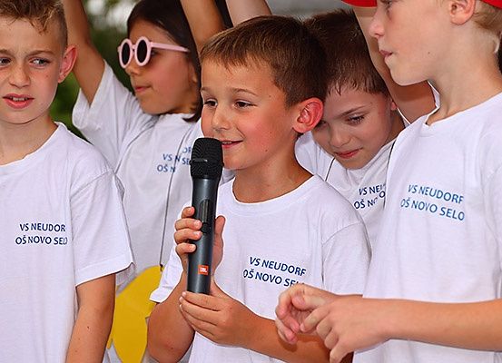Eine Gruppe von Kindern in weißen T-Shirts mit blauem Text, eines hält ein Mikrofon und spricht. Sie stehen draußen.