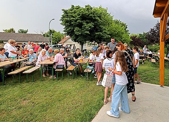 Eine Gruppe von Menschen sitzt auf Picknicktischen in einem Park, einige stehen. Bäume und ein Gebäude sind im Hintergrund.