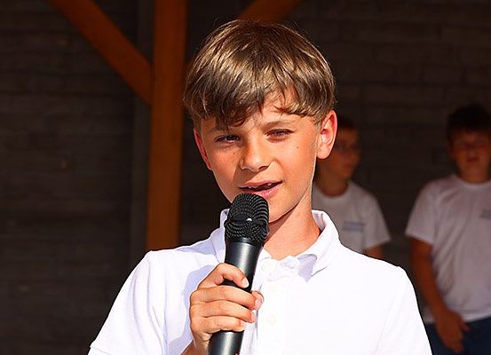 Ein Junge, der ein Mikrofon hält, singt oder spricht, mit zwei Jungen in weißen T-Shirts im Hintergrund.