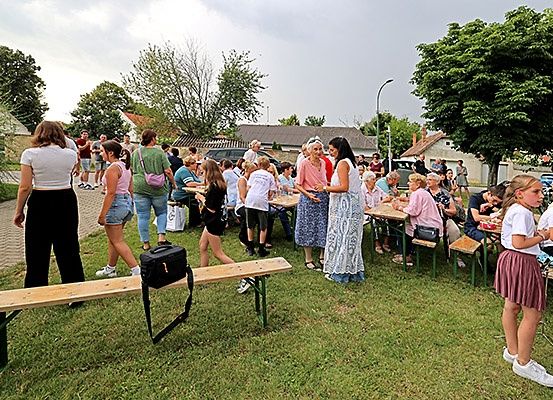 Eine Gruppe von Menschen im Freien auf einer Rasenfläche. Einige sitzen an Tischen, während andere stehen und sich unterhalten. In der Nähe befinden sich Häuser, eine Straßenlaterne und Bäume. Das Wetter ist sonnig.