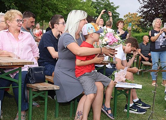 Eine Gruppe von Menschen versammelt sich im Freien, sitzt auf Bänken, wobei eine Frau Blumen hält und ein Junge auf ihrem Schoß sitzt, während andere zusehen und Fotos machen.