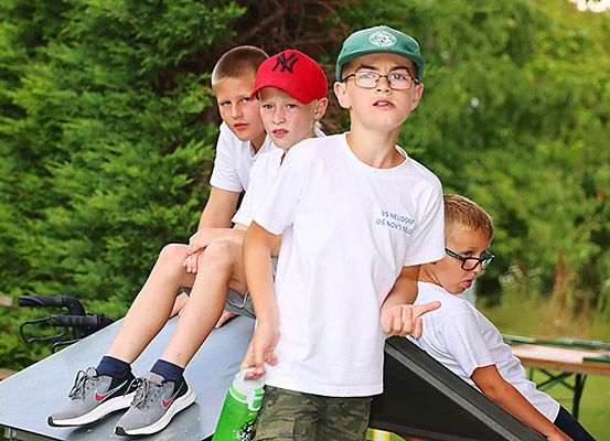 Drei Jungen in weißen T-Shirts sind auf einer Rutsche. Einer hat einen roten Hut, einer einen grünen Hut und einer hält eine Flasche. Hinter ihnen sind Bäume und eine Bank.