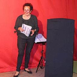 Eine Frau steht vor einem roten Hintergrund und hält eine Broschüre in der Hand. Sie trägt ein gepunktetes Hemd, schwarze Hose und eine Brille. Ein Notenständer steht hinter ihr.