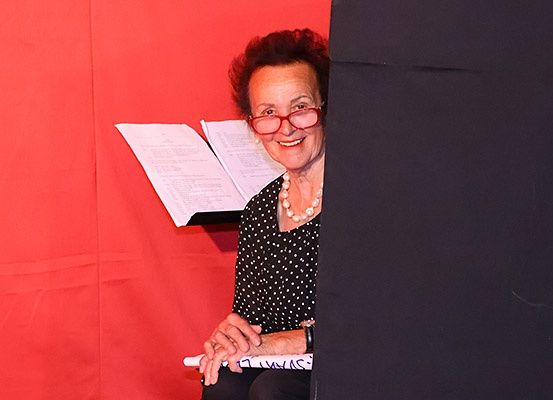 Eine ältere Frau mit Brille und Perlenkette sitzt vor einem roten Hintergrund und hält ein weißes Objekt mit schwarzen Schriftzeichen.