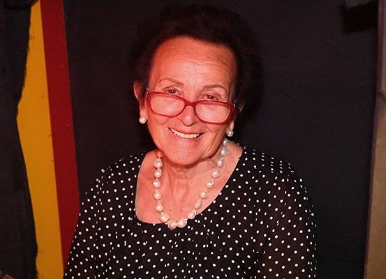 Eine lächelnde ältere Frau mit kurzen Haaren, die rote Brille und eine Perlenkette trägt. Sie steht vor einem dunklen Hintergrund.