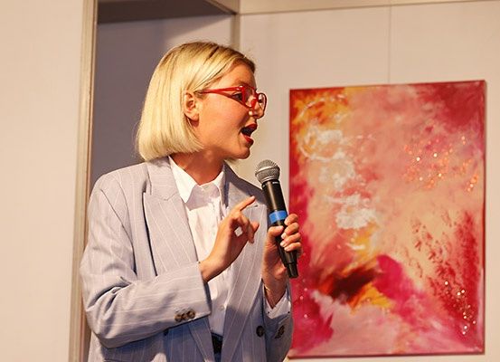 Eine Frau mit Brille hält ein Mikrofon und spricht vor einem farbenfrohen Gemälde.