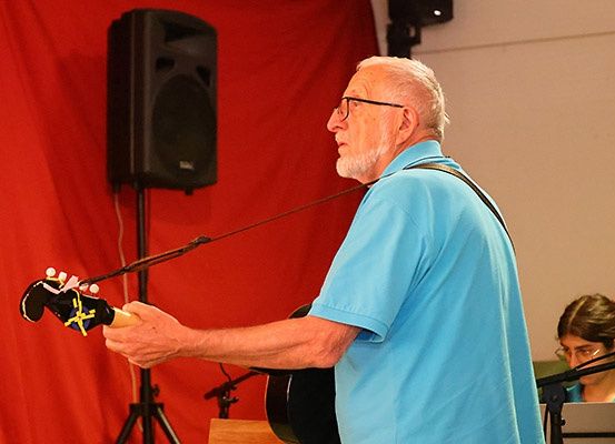Ein älterer Mann mit Brille spielt Gitarre und steht vor einem roten Hintergrund. Hinter ihm spielt eine Frau Klavier.