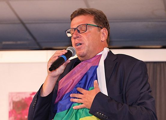 Ein Mann mit Brille und Anzug spricht in ein Mikrofon und hält eine Regenbogenflagge auf der Brust.